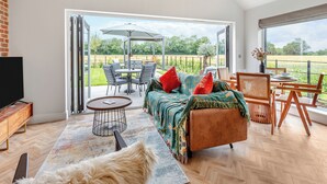 A spacious living area with a sofa, chairs, a TV, and a view of a garden through large windows. - UK47693 - Piglet Cottage (Wimbish)