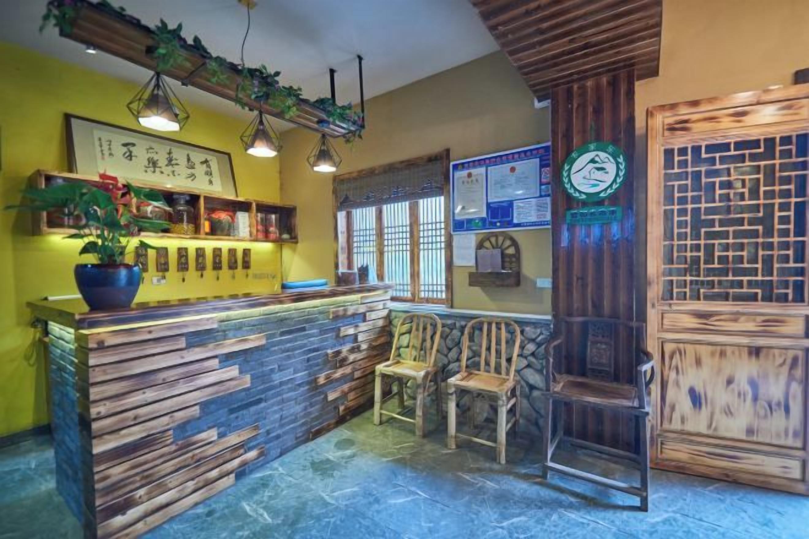 Una zona de recepción con mostrador de madera, dos sillas de madera, una planta en maceta y una pared con un cartel enmarcados y un tablón de anuncios.