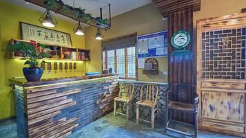 Una zona de recepción con mostrador de madera, dos sillas de madera, una planta en maceta y una pared con un cartel enmarcados y un tablón de anuncios.