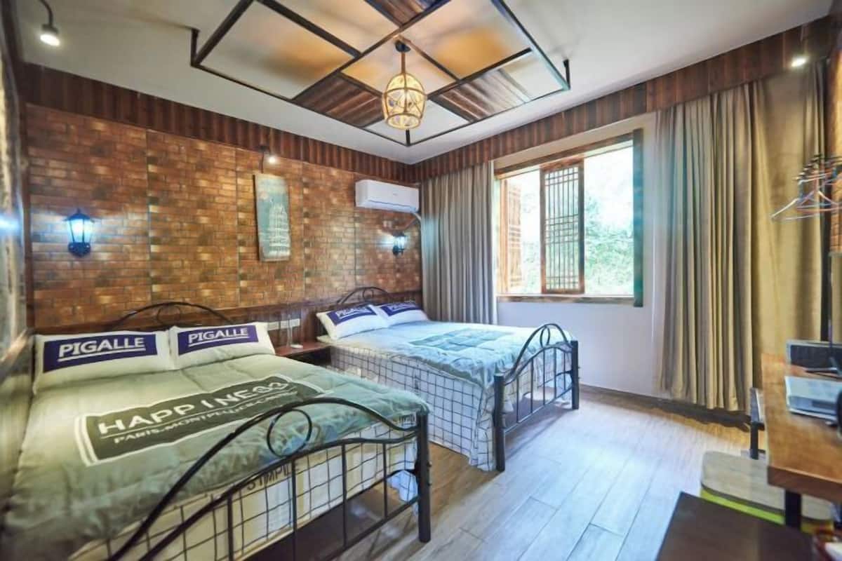 A room with two beds, a brick wall, a window with curtains, a hanging light fixture, and a small table with a chair.