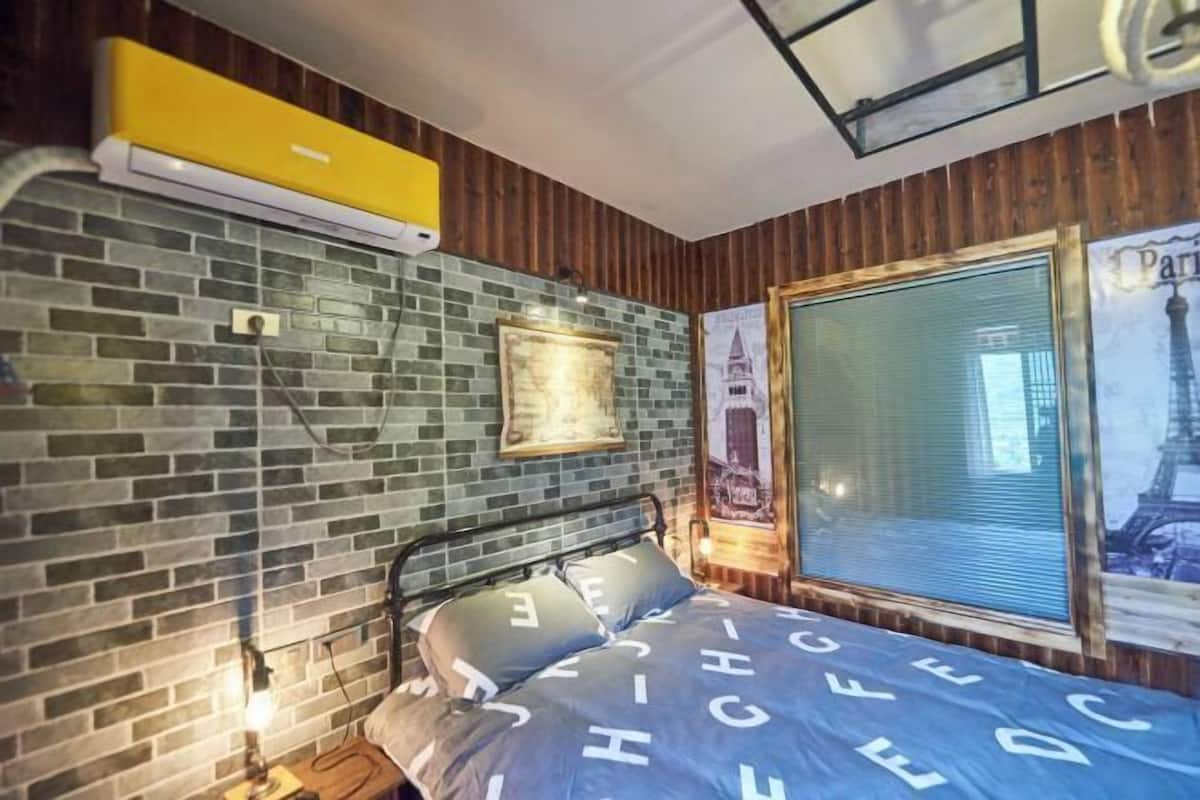 A room with a brick-patterned wall, a bed with a blue bedspread, a window with a view of a building, and a yellow air conditioning unit.