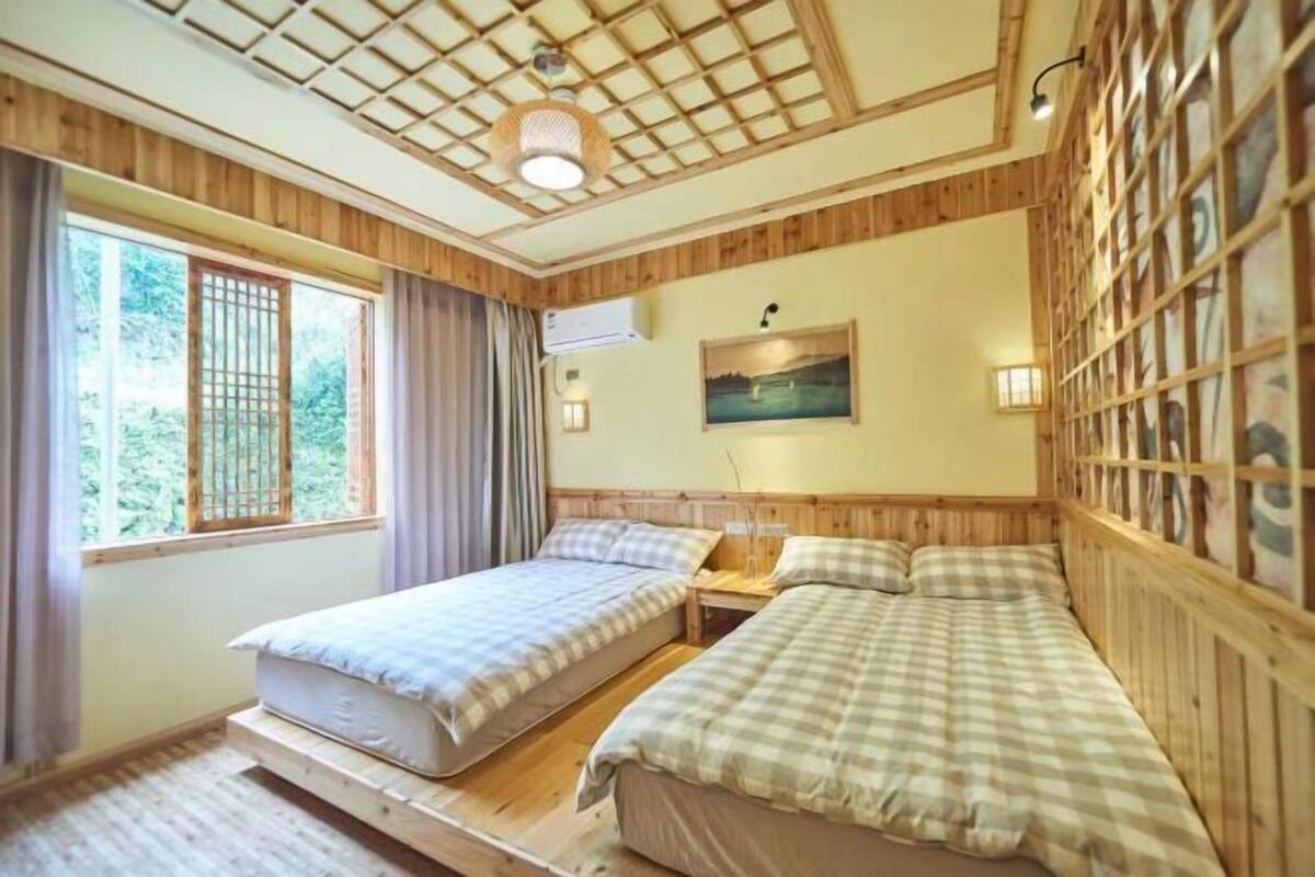 A room with two beds, a window with curtains, a painting on the wall, and a wooden ceiling.