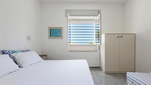Ein Schlafzimmer mit Bett, Kleiderschrank, einem kleinen Tisch, einem Fenster mit Jalousien und einem Bild an der Wand.