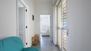 Ein schmaler Flur mit einem petrolfarbenen Stuhl, Holzregalen und einer weißen Tür zu einem Zimmer mit einem Bett.