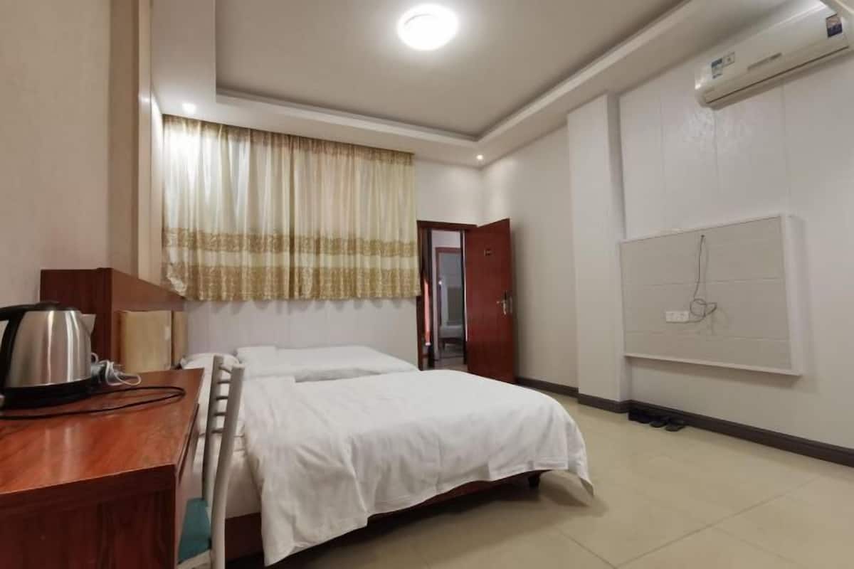 A hotel room with a bed, a desk with a kettle, a window with curtains, an air conditioning unit, and a wall socket.