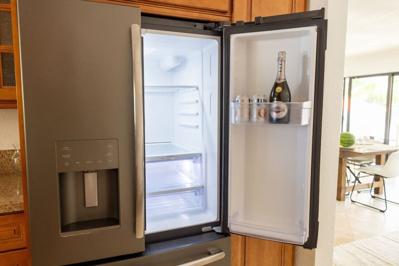 Un refrigerador de acero inoxidable con dispensador de agua, puerta abierta que muestra un interior vacío y una botella de champán en el estante de la puerta.