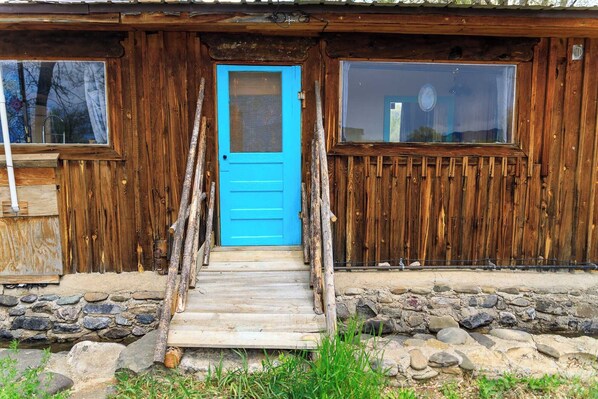 A rustic wooden cabin with a blue door, a stone foundation, and a wooden staircase leading to the entrance.