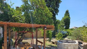 Een rustieke buitenruimte met een houten pergola, picknicktafels, een vuurkorf van natuursteen en een schommel.