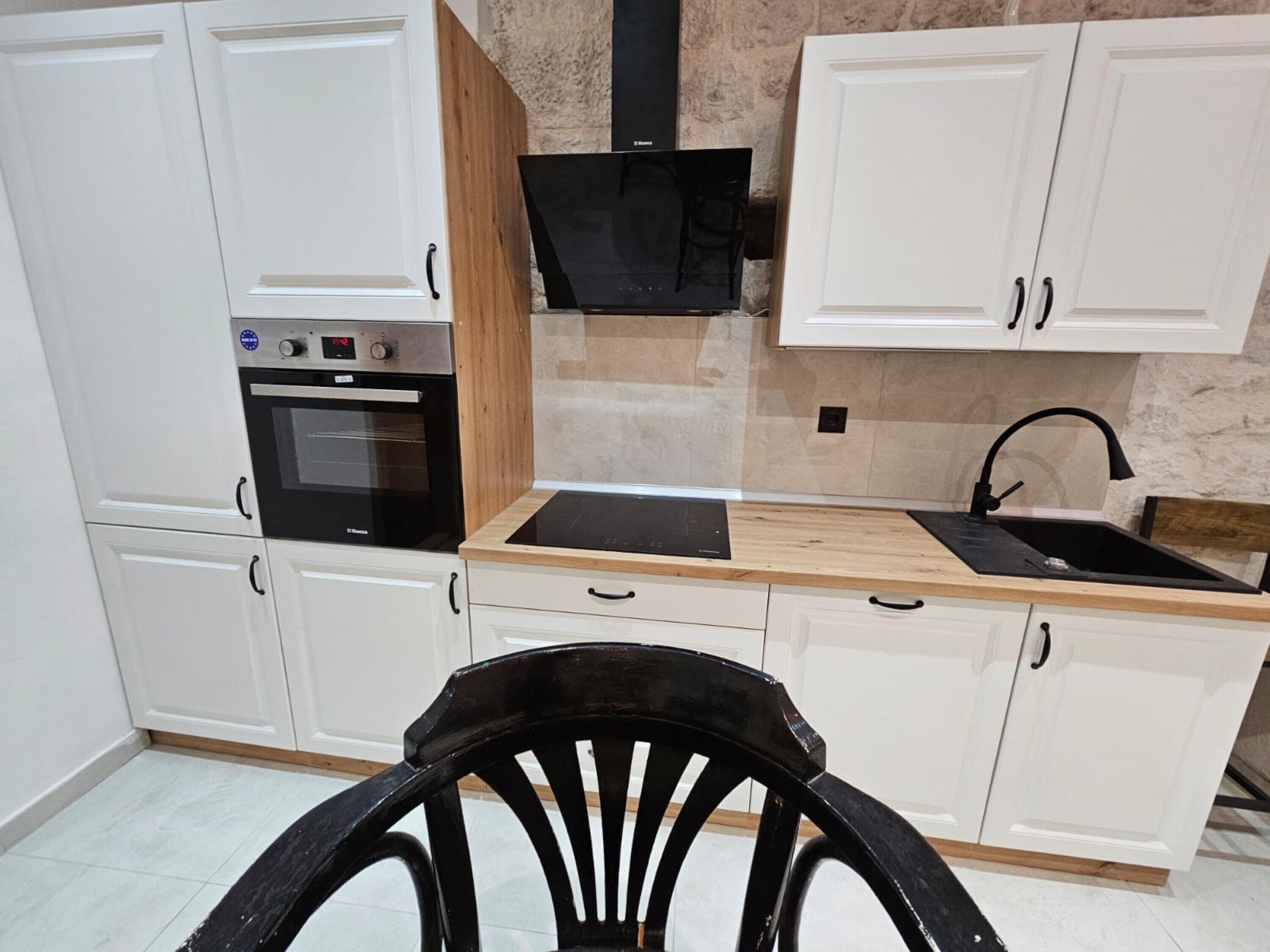Une cuisine avec des meubles blancs, un four noir, un plan de travail en bois et une chaise noire.