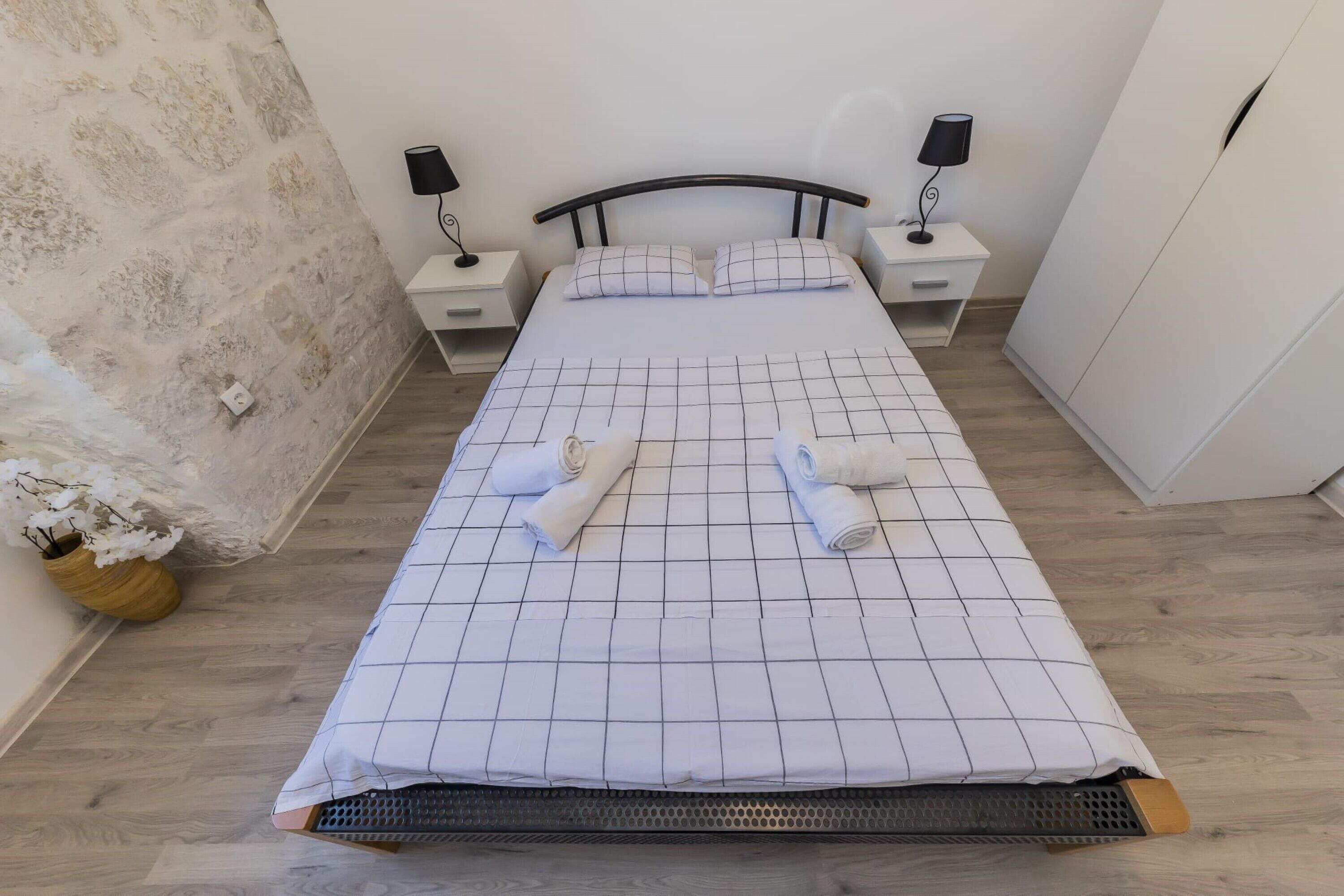 Een netjes opgemaakt bed met een bedspreider met ruitmotief, twee nachtkastjes met lampen, een houten nachtkastje en een vaas met witte bloemen.