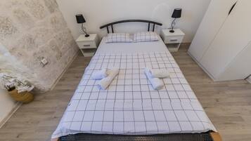 Een netjes opgemaakt bed met een bedspreider met ruitmotief, twee nachtkastjes met lampen, een houten nachtkastje en een vaas met witte bloemen.