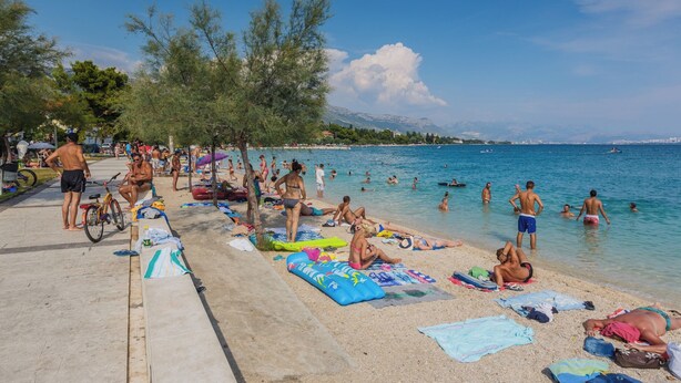 Een strand met mensen die zonnebaden en zwemmen, een betonpad en een helderblauwe lucht.