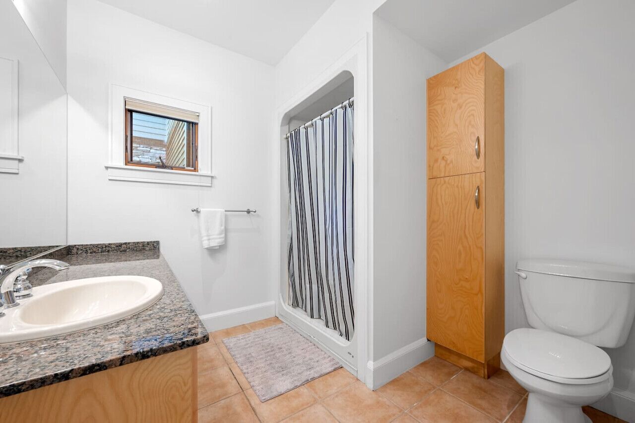 A bathroom with a granite countertop sink, a window with blinds, a shower enclosure with a striped curtain, a wooden cabinet, and a toilet.