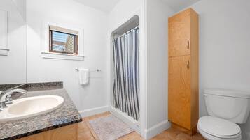 A bathroom with a granite countertop sink, a window with blinds, a shower enclosure with a striped curtain, a wooden cabinet, and a toilet.