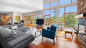 Ein geräumiges Wohnzimmer mit einem großen grauen Ecksofa, einem blauen Sessel und einem Glastisch. Ein Fernseher steht auf einem Lowboard, es gibt einen Kamin und große Fenster mit Blick auf Bäume und einen See.