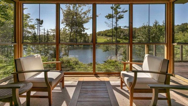 Eine Holzterrasse mit zwei Stühlen und einem Tisch, mit Blick auf einen See und einen Wald.