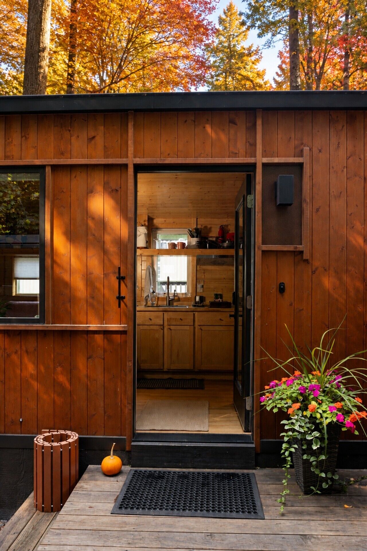 A wooden cabin with a kitchen area visible through an open door, a small outdoor seating area with a pumpkin, and a planter with flowers.