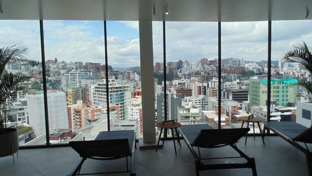Quarto com grandes janelas com vista para a cidade, uma mesa pequena e duas cadeiras.