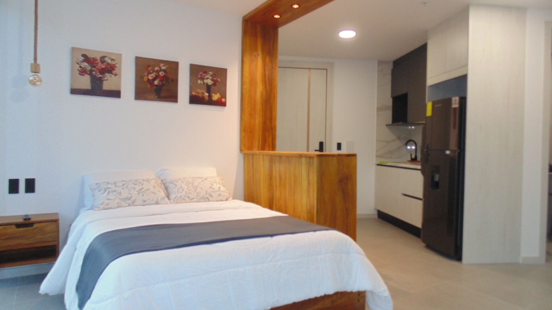 A bedroom with a bed, a wooden headboard, and a kitchenette with a refrigerator.