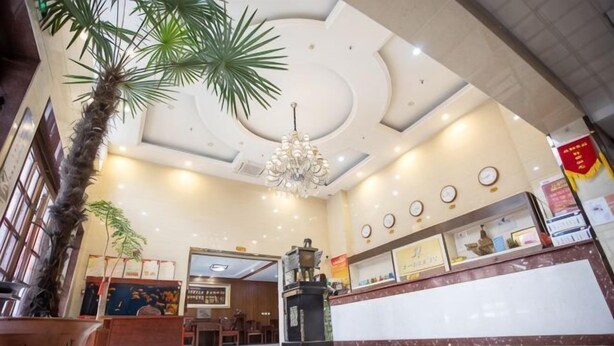 A lobby with a reception desk, a chandelier, a palm tree, and a large mirror. - Youyichun Business Hotel (Zhao County)
