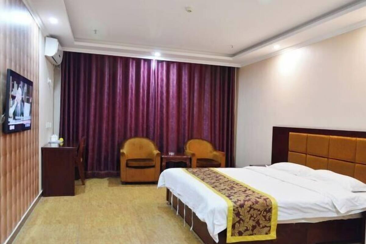Hotellihuone, jossa on sänky, kaksi tuolia, pieni pöytä, televisio ja verho.
