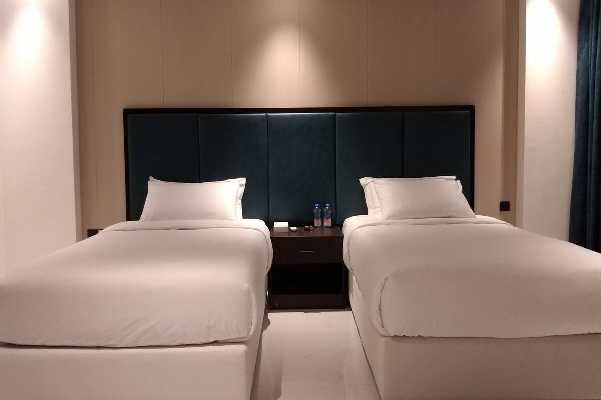 A hotel room with two single beds, a dark headboard, and a nightstand with water bottles.
