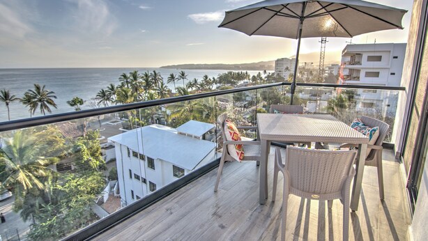 A balcony with a table, chairs, and an umbrella overlooking a coastal view with palm trees and buildings. - Oceanview Penthouse Condo in Sunny Bucerias (Bucerias)