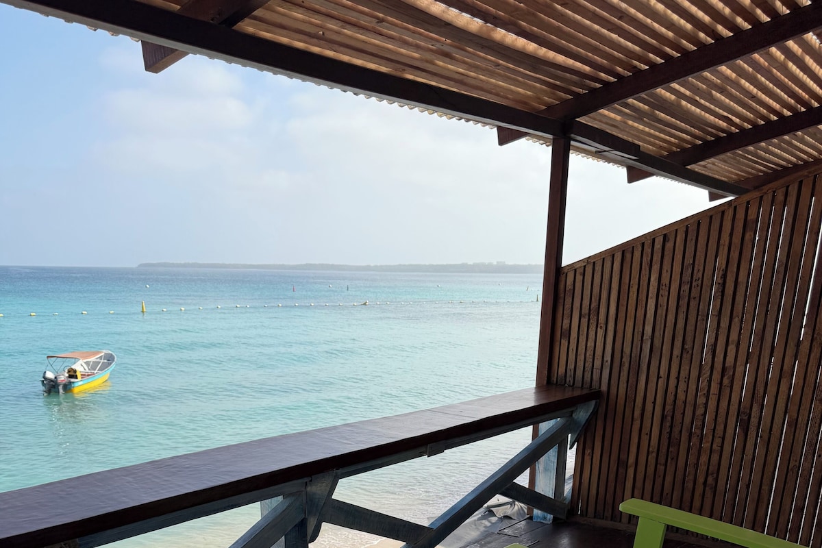 A wooden deck with a railing overlooking a beach, a small boat on the water, and colorful outdoor furniture.