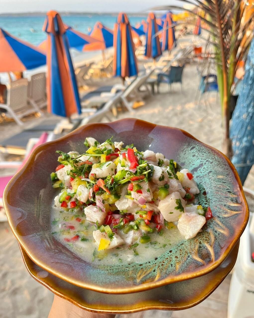 A bowl of seafood salad on a beach with umbrellas and lounge chairs in the background.