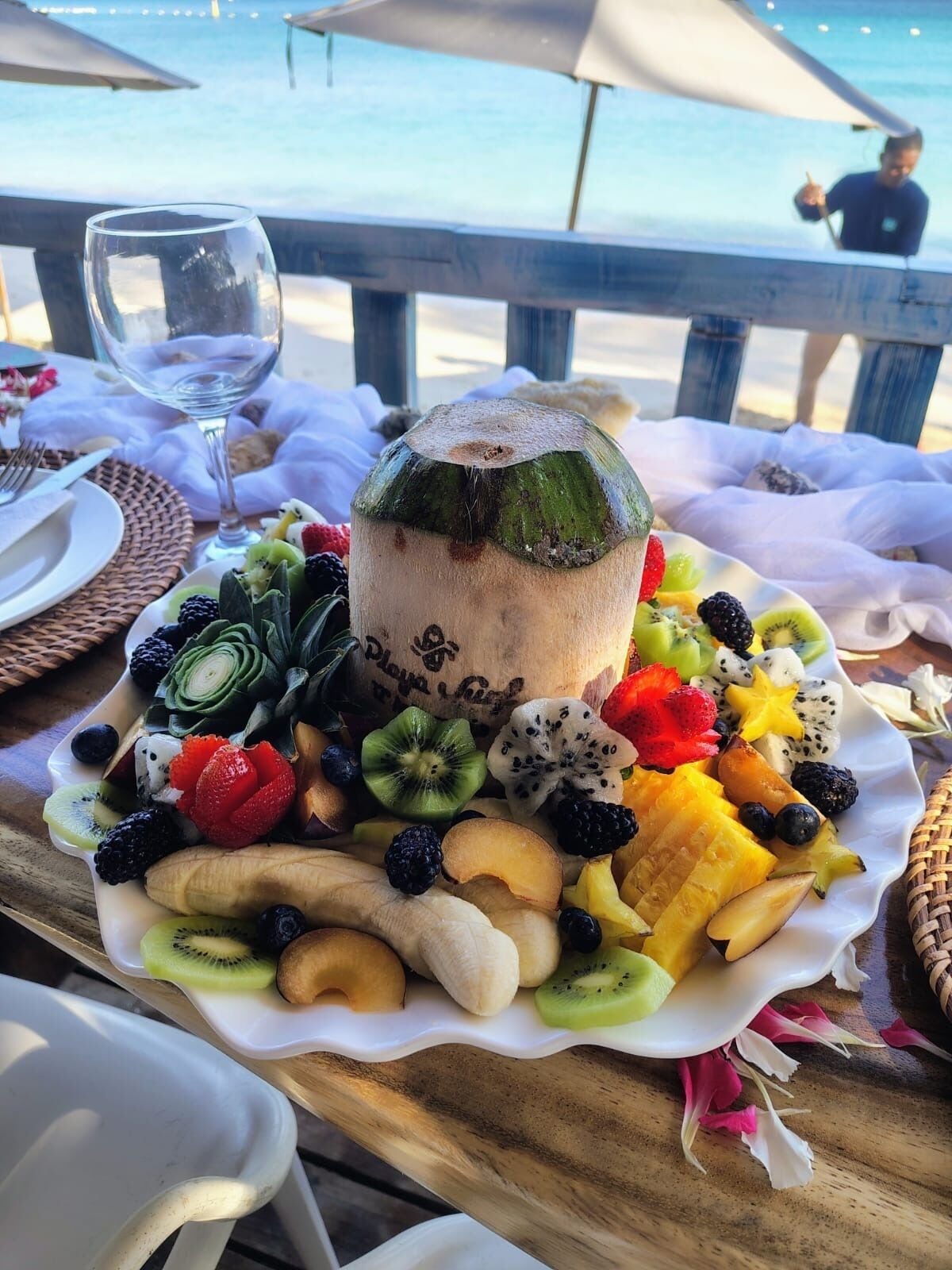 Ein Tisch mit einer Obstplatte, einem Glas Wasser und einem Kokosnussgetränk am Strand.