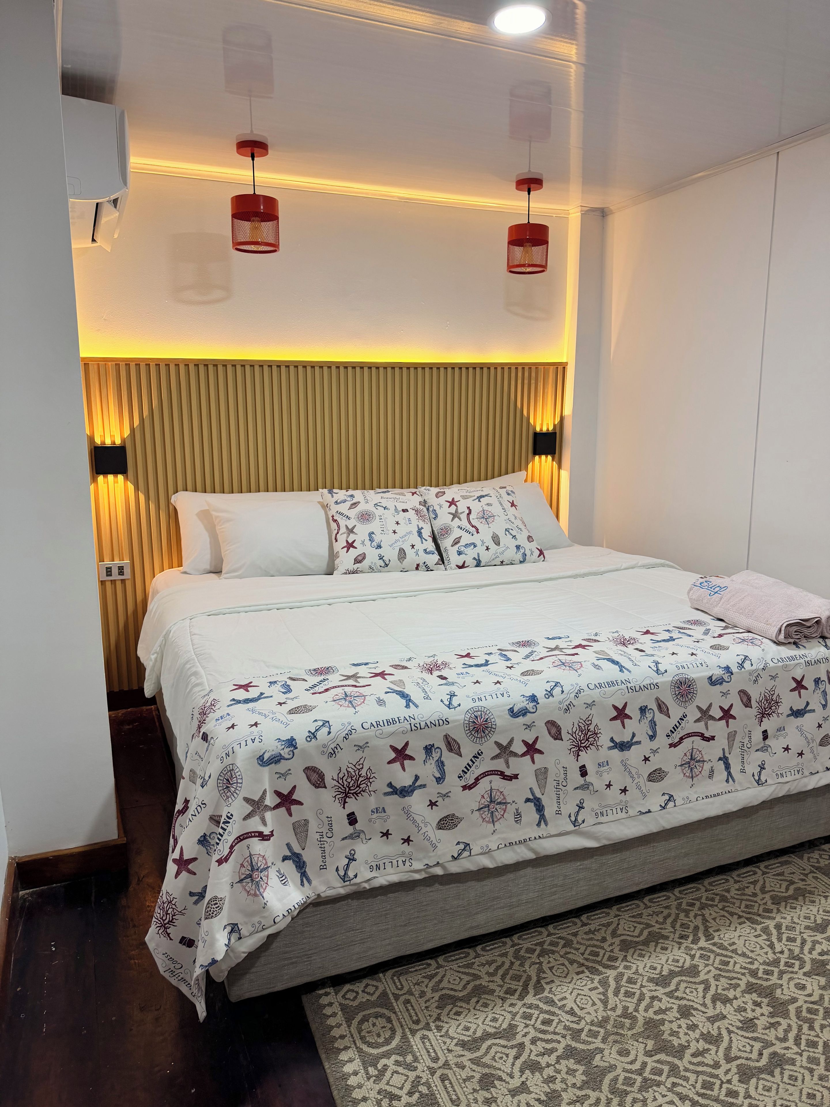 A hotel room with a bed, a headboard, a wall-mounted air conditioner, and a patterned rug.