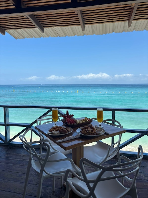 Une table dressée avec de la nourriture et des boissons, donnant sur une plage à l’eau cristalline et une terrasse en bois.