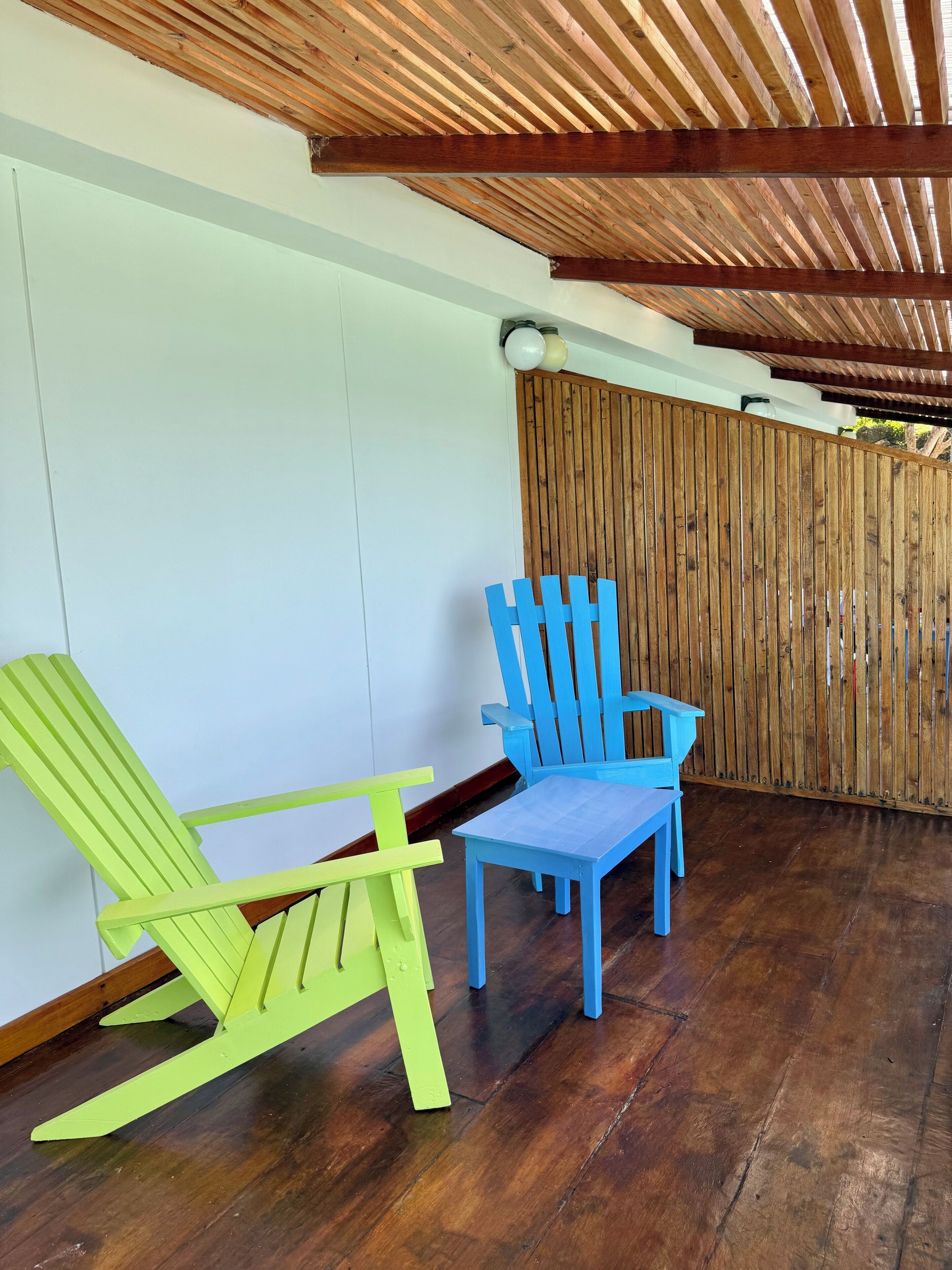 Een overdekt terras met twee stoelen, een groene en een blauwe, en een houten hek op de achtergrond.