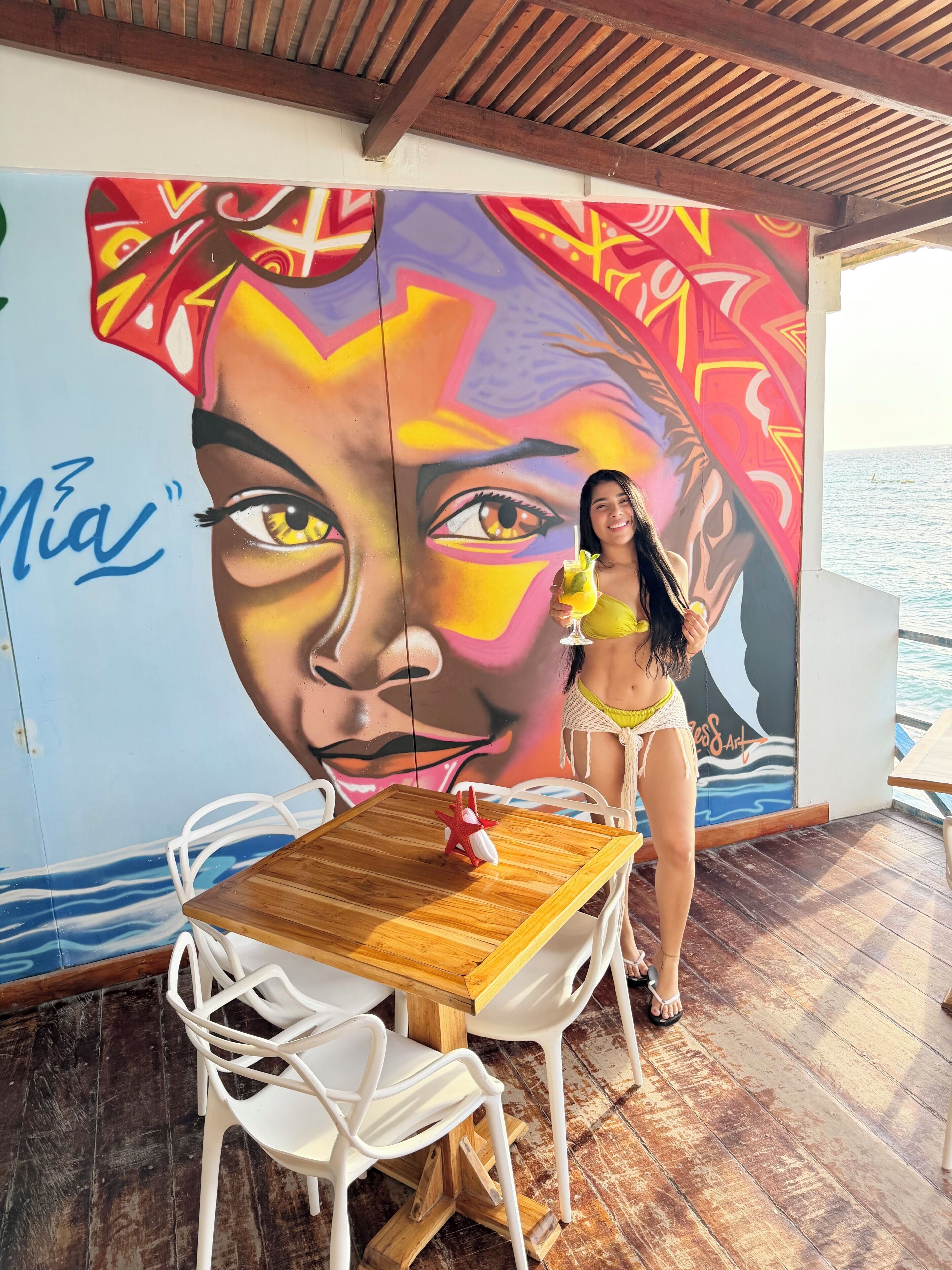 Une personne se tient près d’une table, devant une fresque murale, avec des chaises blanches et une table en bois.