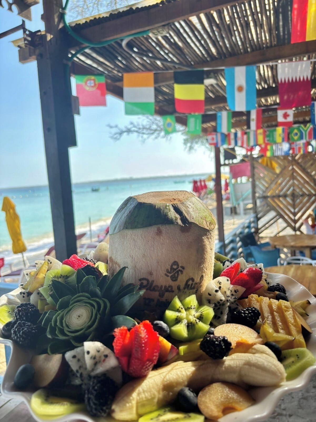 A fruit bowl with various fruits and a coconut on a table, with a beach and flags in the background.
