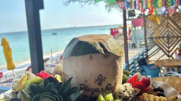 A fruit bowl with various fruits and a coconut on a table, with a beach and flags in the background.