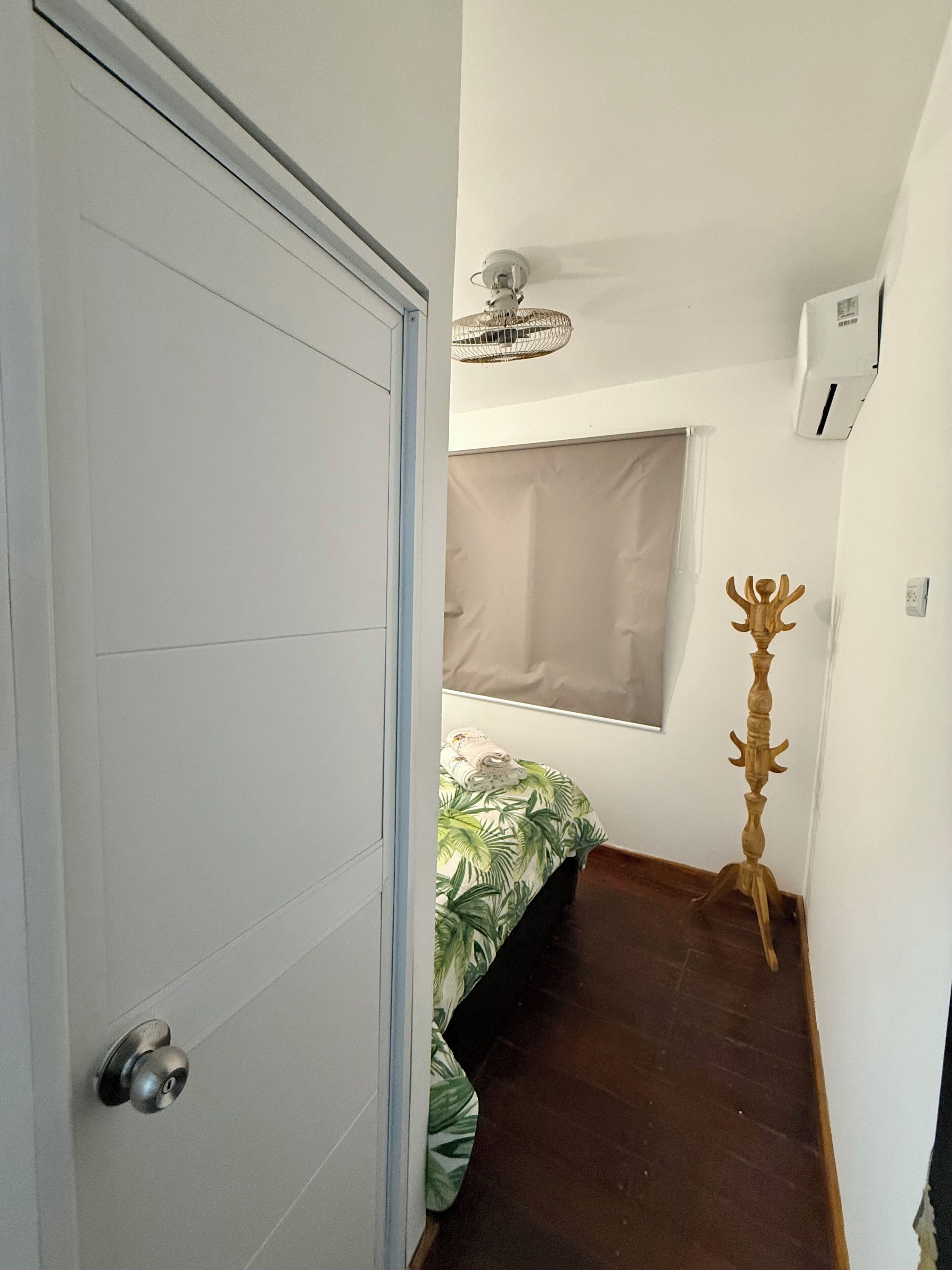 Ein schmales Entree mit einer geschlossenen Tür, einem Bett mit einem Bettbezug mit grünem Blattmuster, einem Fenster mit einer beigen Jalousie, einer Stehlampe aus Holz und einer Klimaanlage an der Wand.