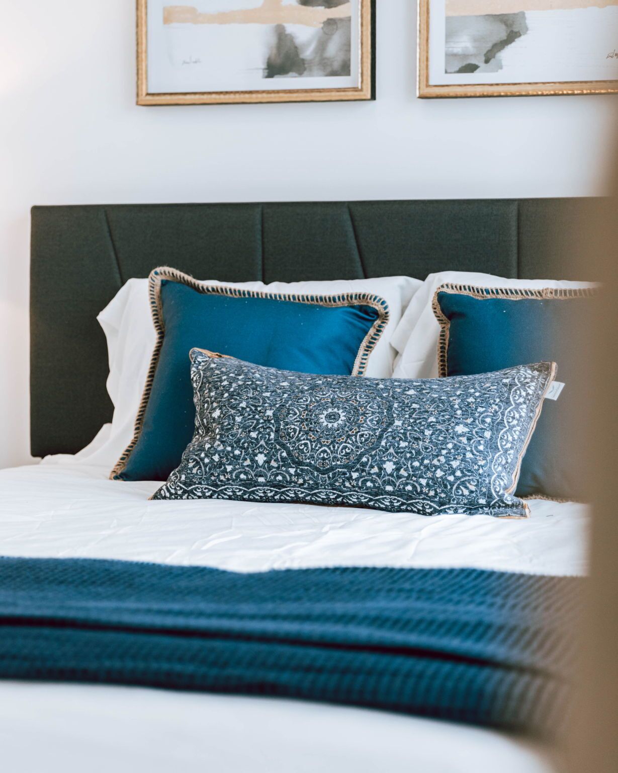 A neatly made bed with a patterned pillow and two plain pillows, a grey upholstered headboard, and two framed artworks on the wall.