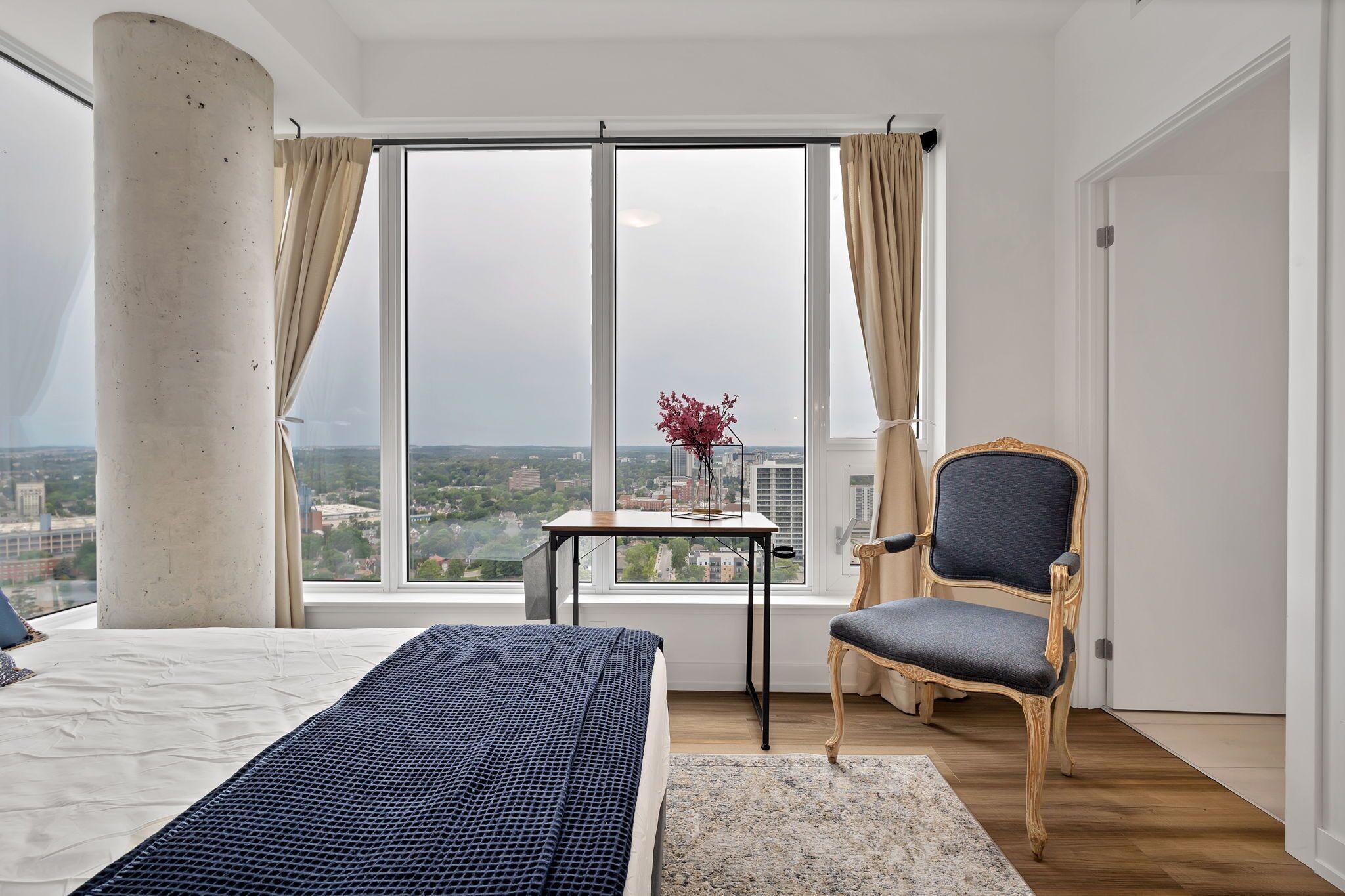 A bedroom with a large window, a bed, a chair, and a small table with a vase.