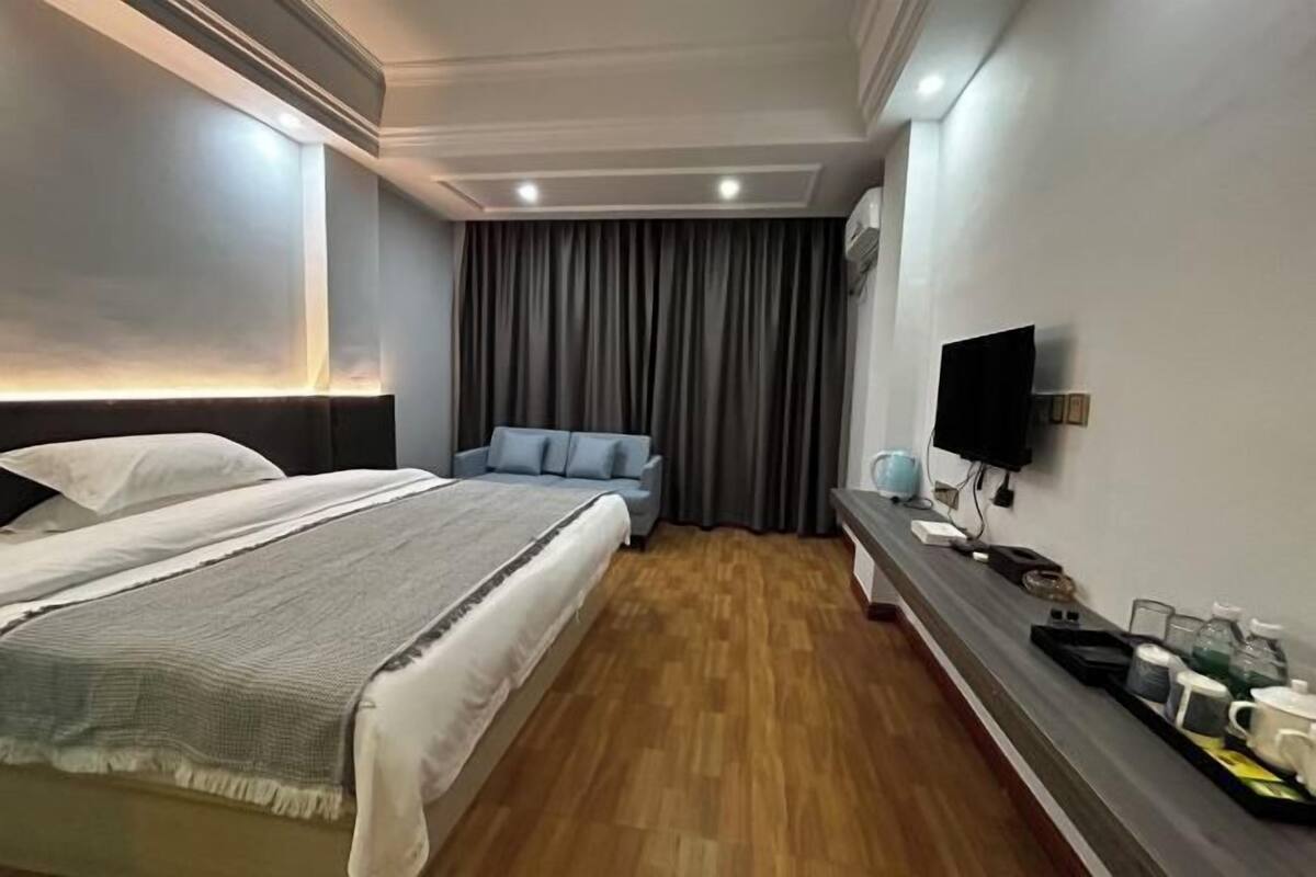 A hotel room with a large bed, a smaller sofa, a TV, and a bedside table with a tray of items.