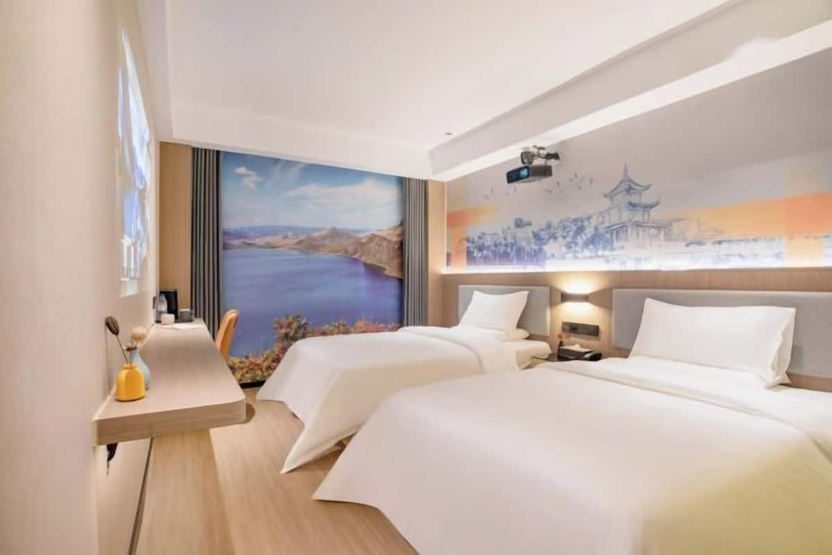 A hotel room with two beds, a desk with a vase, and a mural of a landscape.