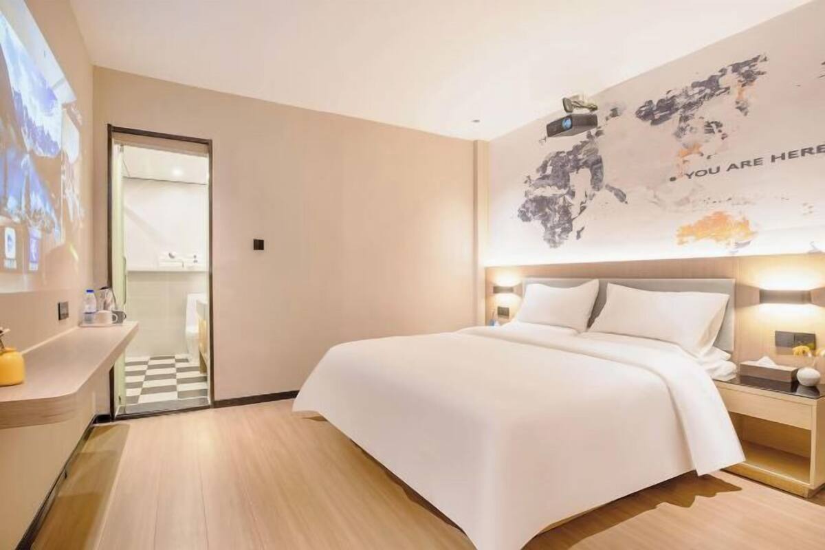 A hotel room with a bed, a desk, a nightstand, a wall-mounted TV, and a map on the wall.