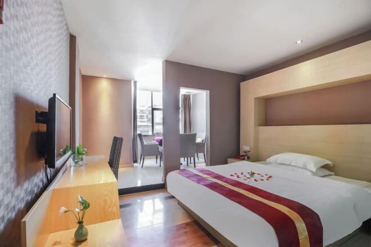 A hotel room with a large bed, a TV, a desk with a vase, and a view of the outside.