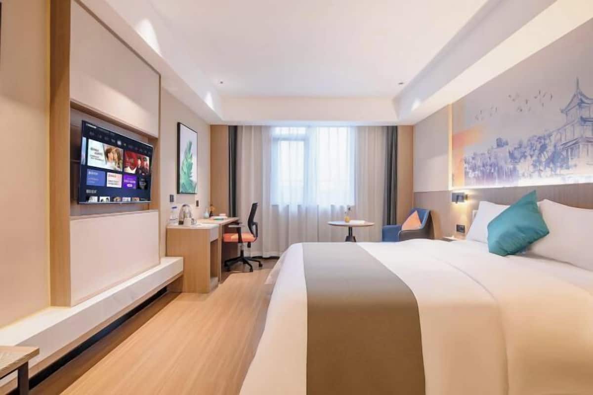A hotel room with a bed, desk, chair, TV, and artwork on the wall.
