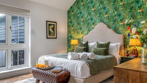 A bedroom with a bed, pillows, a bedside table with a bottle and glasses, a window, and a wall with a floral pattern.