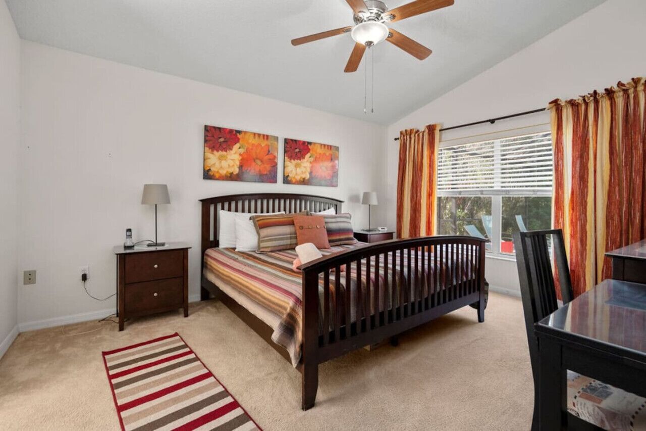 Une chambre à coucher comprenant un lit, des tables de chevet, un ventilateur de plafond, une fenêtre avec des rideaux et une vue sur l’extérieur.