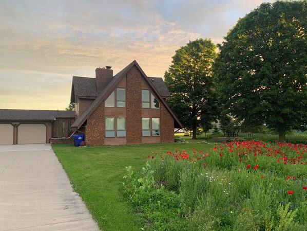 A two-story house with a brick facade, large windows, and a visible garage. - Our Thunder Moon Farm Stay- sleeps 7, framed by wildflowers. 4 miles to GB. (Oneida)