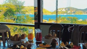A breakfast buffet with various pastries, fruits, and beverages, set against a scenic view of a beach and hills. - Pousada Encanto de Buzios (Búzios)