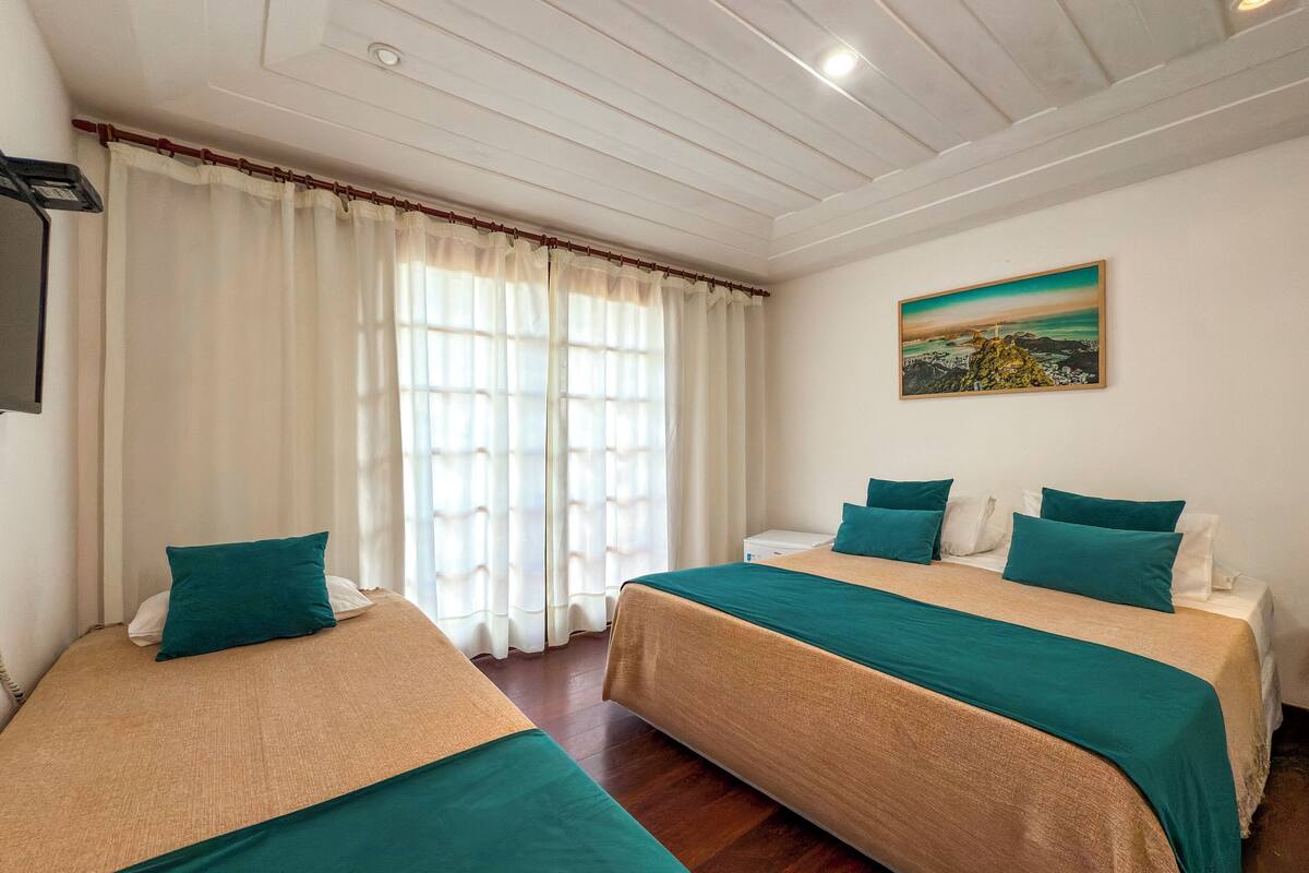 Habitación de hotel con dos camas, cada una con ropa de cama color aguamarina, un ventanal con cortinas blancas y un cuadro de una escena costera en la pared.