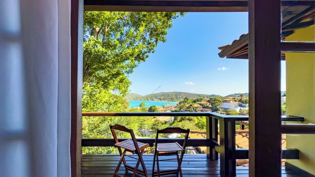 A wooden deck with two chairs and a table, overlooking a scenic view of a coastal area with houses and greenery. - Pousada Encanto de Buzios (Búzios)
